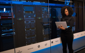 A woman with curly hair stands in a data center, using a laptop to interact with the server equipment behind her, suggesting the management of complex IT infrastructure.