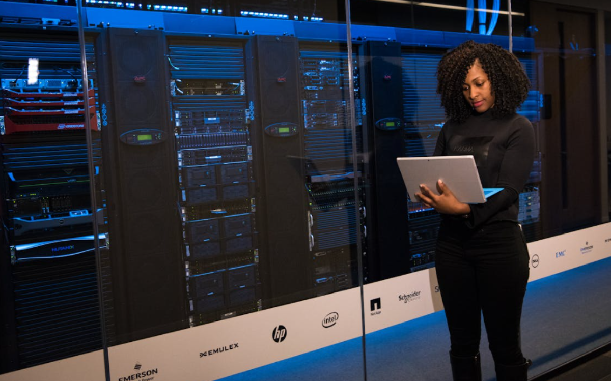 A woman with curly hair stands in a data center, using a laptop to interact with the server equipment behind her, suggesting the management of complex IT infrastructure.