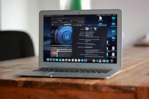 Laptop computer on a wooden desk, screen showing Mac system preferences and a cybersecurity webpage, with application icons in the dock
