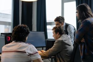 Software developers or IT workers gathered around a desktop computer, analyzing something on the screen in a bright, minimalist workspace.