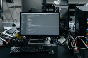 Coding workspace with desktop computer, monitor showing software development code, and various computer components and wires in a cluttered tech setup