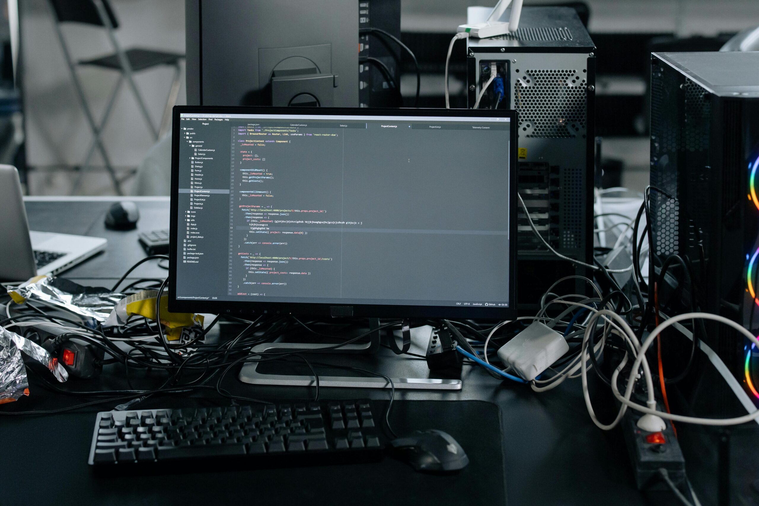 Coding workspace with desktop computer, monitor showing software development code, and various computer components and wires in a cluttered tech setup