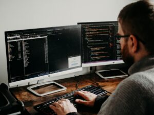 Software developer working on code, viewed from behind, with two computer monitors displaying programming interfaces and dark coding screens