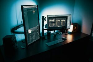 Professional dual-monitor workstation with blue ambient lighting, showing digital interface screens, keyboard, speakers, and computer accessories on a dark desk