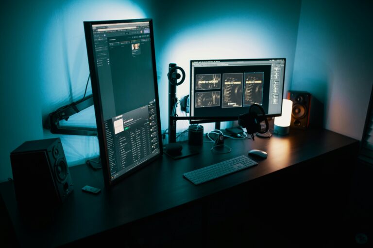 Professional dual-monitor workstation with blue ambient lighting, showing digital interface screens, keyboard, speakers, and computer accessories on a dark desk