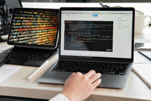 Software development workspace with a laptop showing vibrant, multicolored source code and another laptop displaying Java code in a development environment, with a hand positioned on the keyboard.