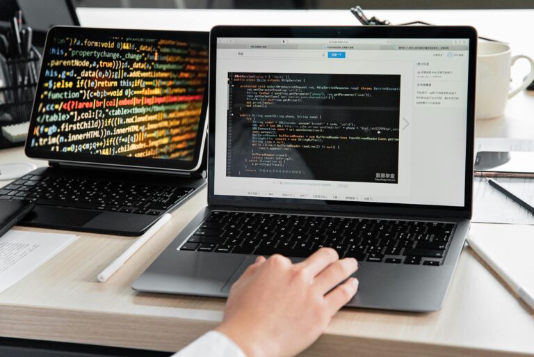 Software development workspace with a laptop showing vibrant, multicolored source code and another laptop displaying Java code in a development environment, with a hand positioned on the keyboard.