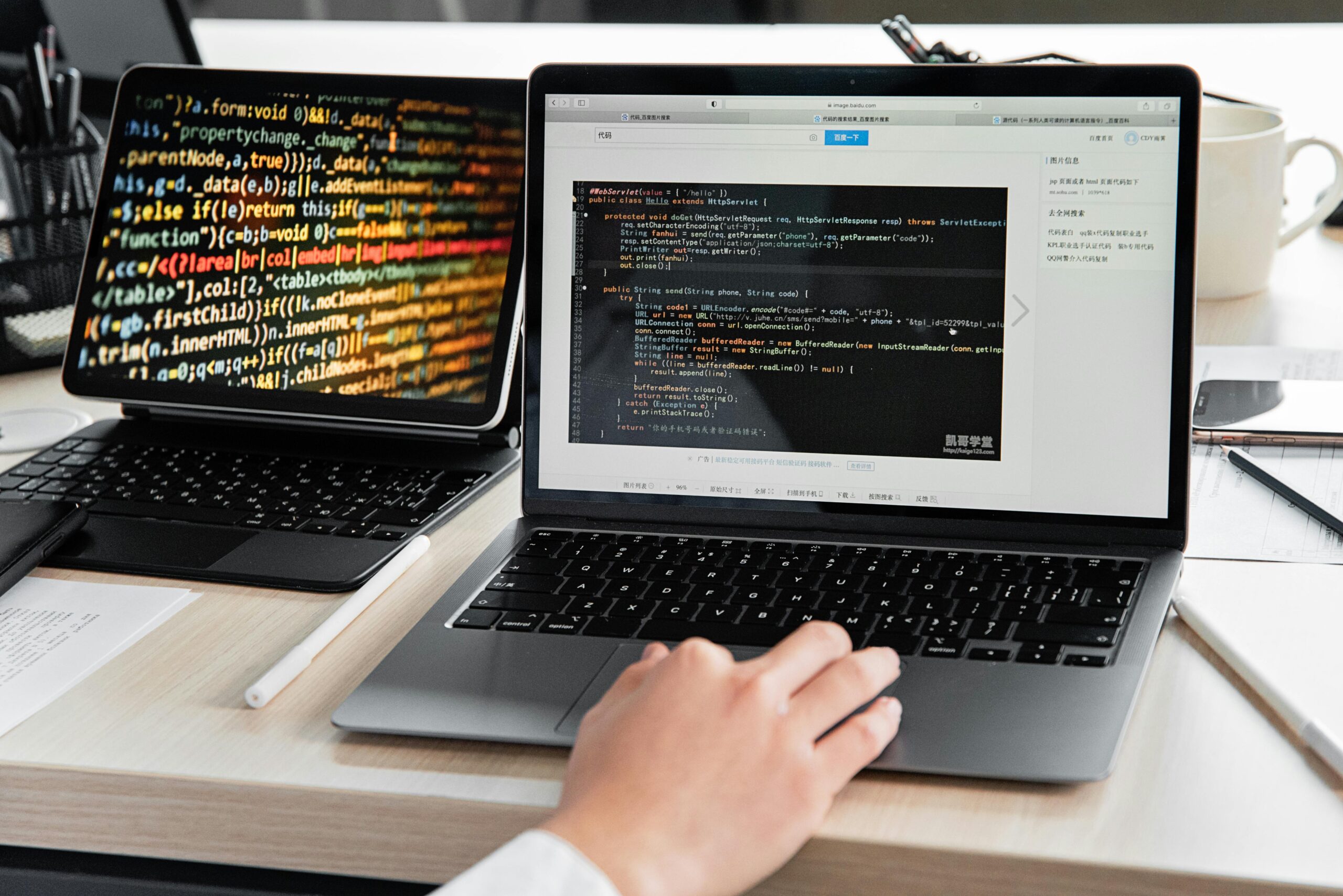 Software development workspace with a laptop showing vibrant, multicolored source code and another laptop displaying Java code in a development environment, with a hand positioned on the keyboard.