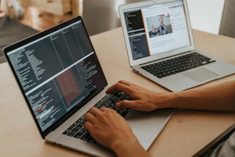Software development workspace featuring two silver laptops, with source code displayed on dark screens and hands positioned on a keyboard