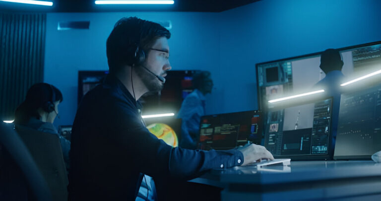 Data center or mission control scene with professionals seated at computer stations, illuminated by cool blue overhead lights and display screens