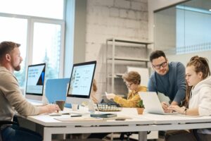 A diverse team of professionals engaged in a meeting or project discussion, using laptops, tablets, and paper materials to coordinate their work.