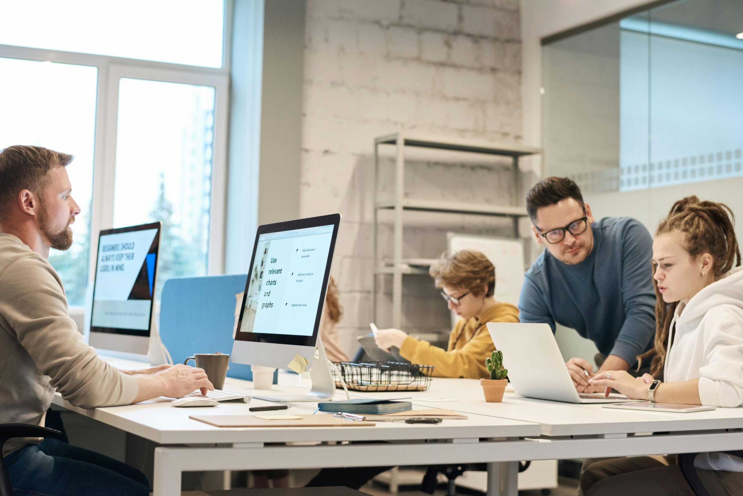 A diverse team of professionals engaged in a meeting or project discussion, using laptops, tablets, and paper materials to coordinate their work.