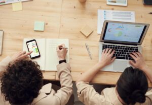 Overhead view of a desk workspace showing technology tools like a laptop and smartphone, along with analog materials like notebooks and sticky notes, demonstrating a blended approach to productivity.