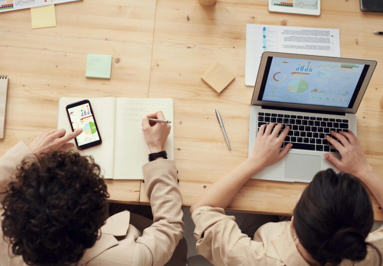 Overhead view of a desk workspace showing technology tools like a laptop and smartphone, along with analog materials like notebooks and sticky notes, demonstrating a blended approach to productivity.
