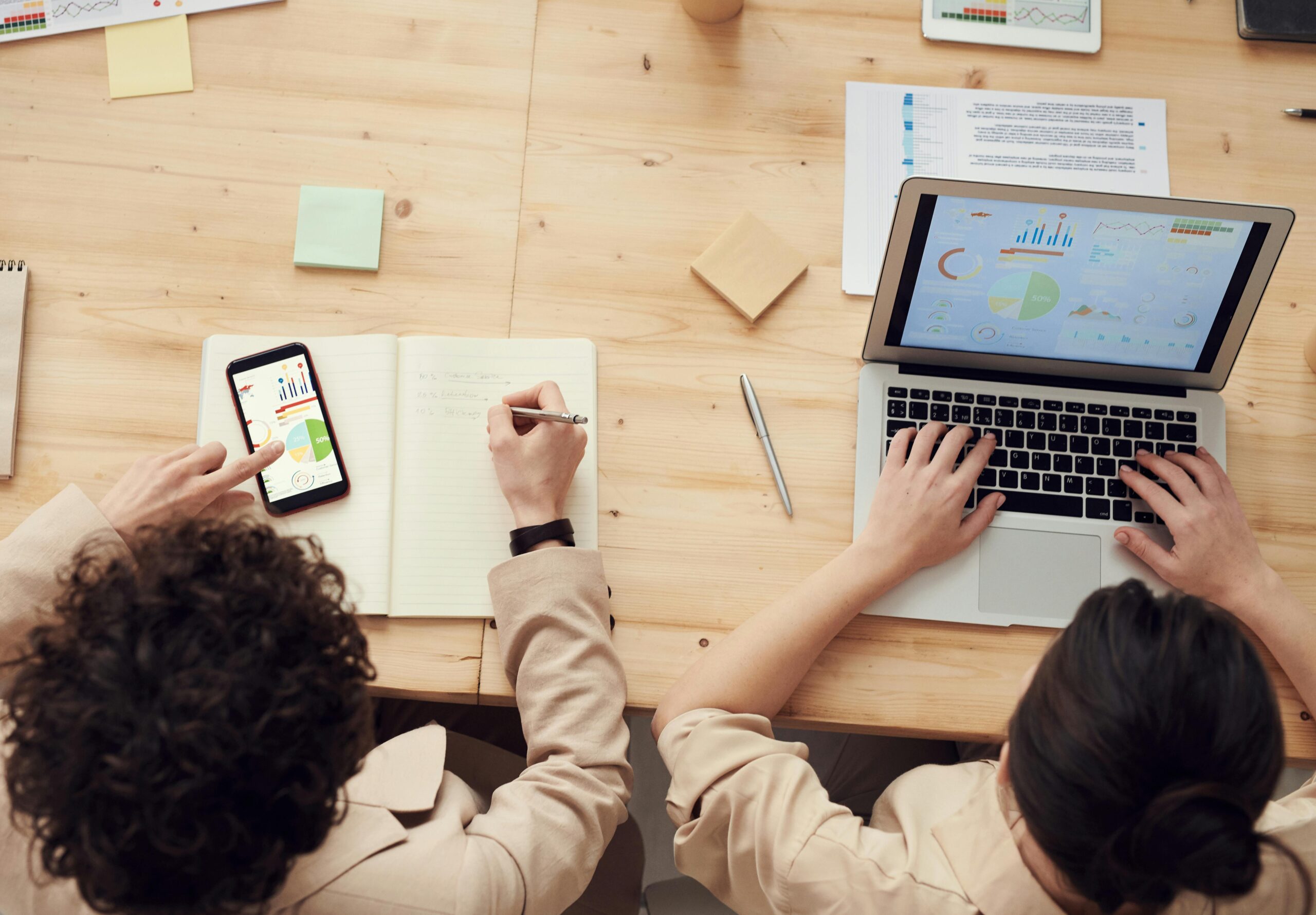 Overhead view of a desk workspace showing technology tools like a laptop and smartphone, along with analog materials like notebooks and sticky notes, demonstrating a blended approach to productivity.
