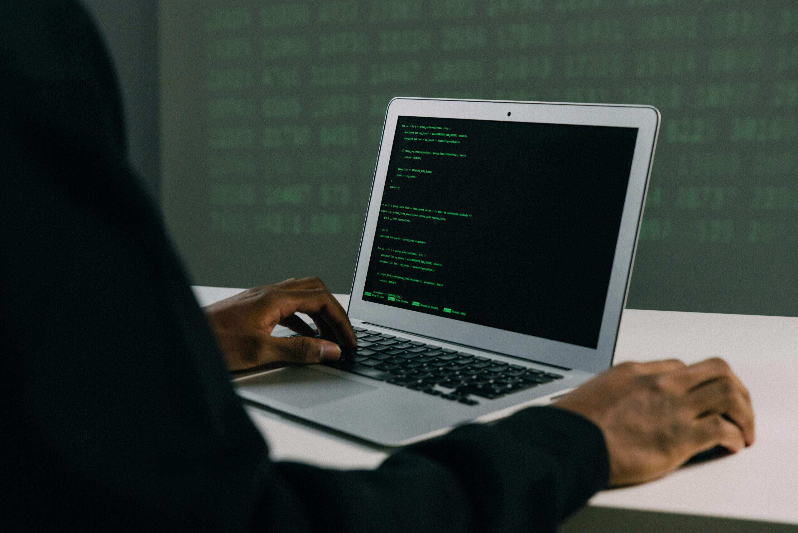 Cybersecurity or software development scene: hands typing on a silver laptop with green code lines visible on the display, set against a blurred green matrix-like background.