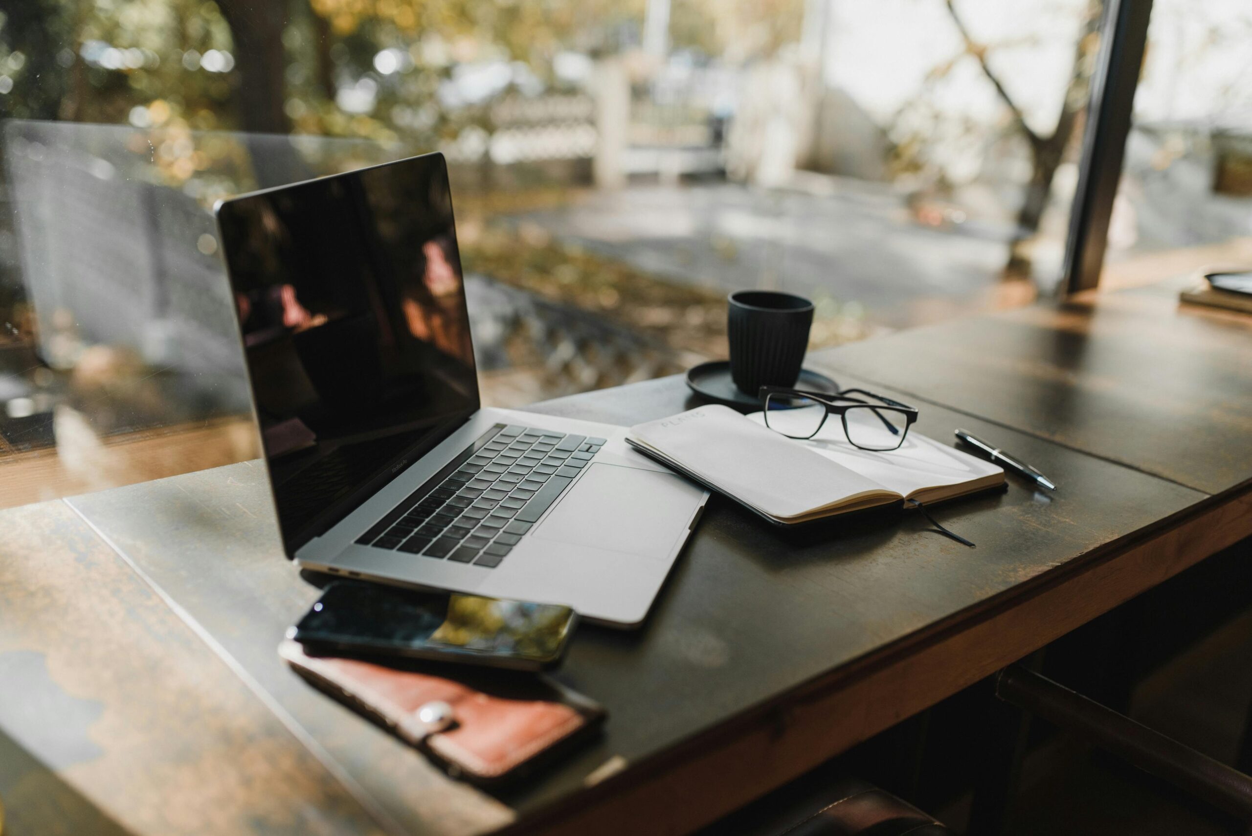 Modern home office setup with a laptop and open notebook on a wooden desk, surrounded by natural light and trees outside the window.