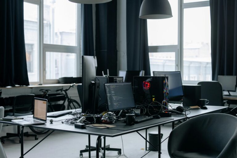Coding Retreat: A dark, moody office environment with large windows, showcasing a desk crowded with multiple screens, keyboards, and peripherals, suggesting a space for intensive coding and software development.