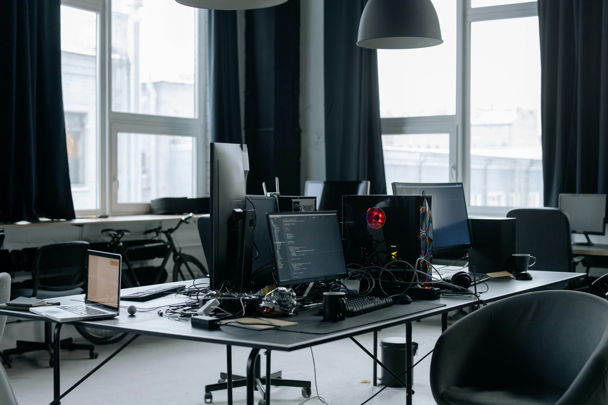 Coding Retreat: A dark, moody office environment with large windows, showcasing a desk crowded with multiple screens, keyboards, and peripherals, suggesting a space for intensive coding and software development.