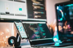 A technology-focused workstation with a smartphone, laptop, and desktop monitors showcasing programming interfaces and coding environments, creating an atmosphere of digital innovation and technological advancement.