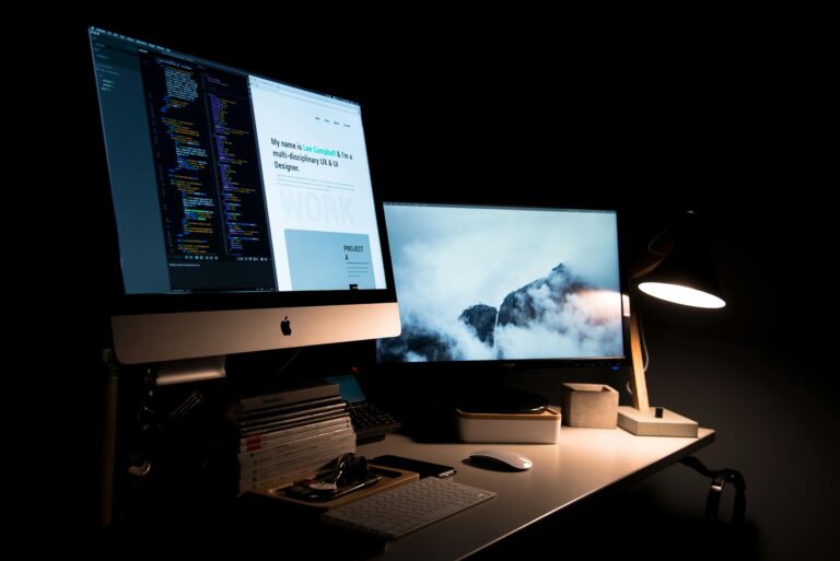 A desk setup with an iMac computer displaying a personal profile on one screen and a breathtaking mountain scene on the other. The workspace is surrounded by various organizational items, suggesting a productive and creative work atmosphere.