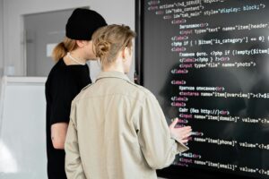 A classroom or workspace setting, with two individuals engaged in a discussion or analysis of the code displayed on the blackboard.