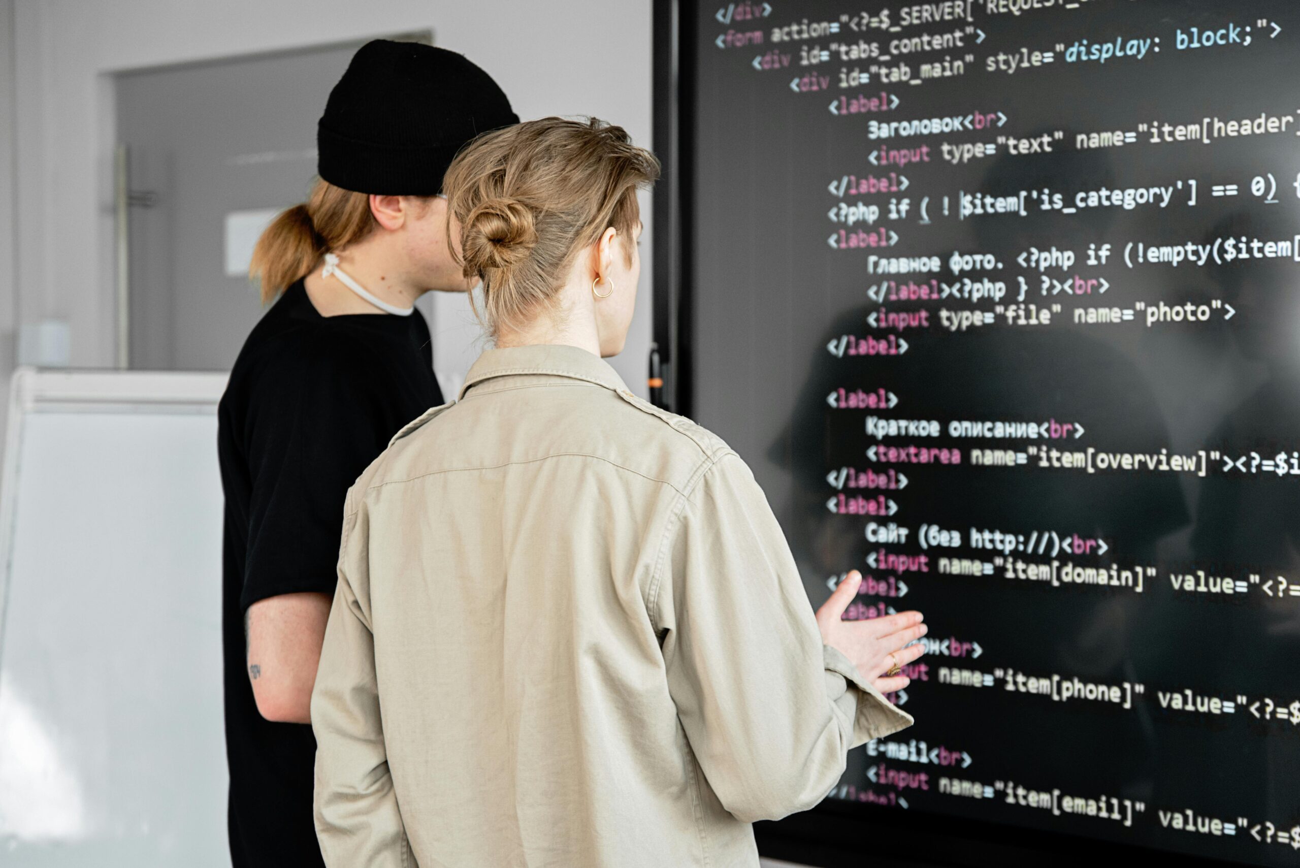 A classroom or workspace setting, with two individuals engaged in a discussion or analysis of the code displayed on the blackboard.
