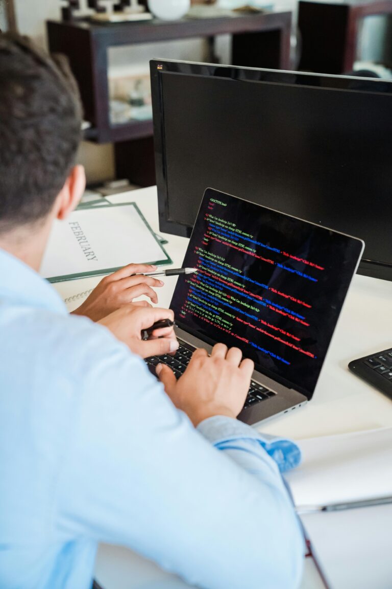 A close-up view of a person's hands typing on a laptop keyboard, with the laptop screen displaying lines of code in various colors.