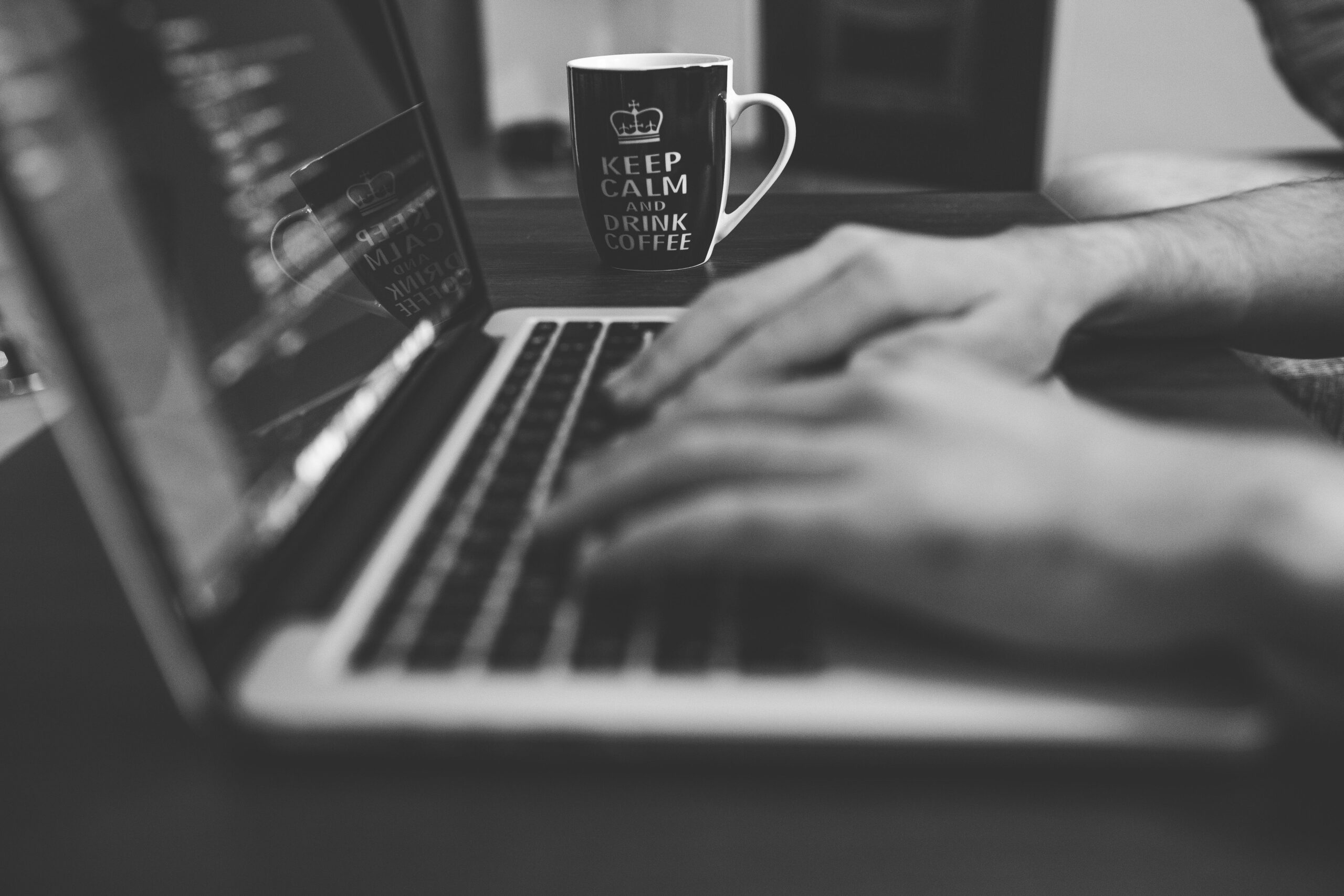 A monochrome image showing hands working on a laptop, with a coffee mug prominently displayed.