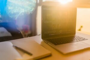 Open laptop displaying photo editing software beside a notebook and pen, bathed in bright, golden sunlight.