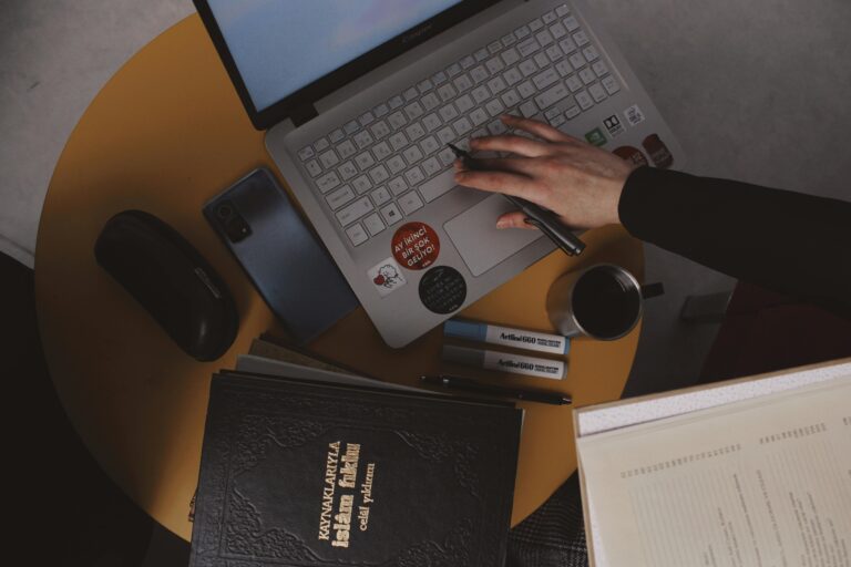 A hand typing on a sticker-covered laptop at a cozy desk setup with coffee, a phone, and study tools, blending tech and tradition.