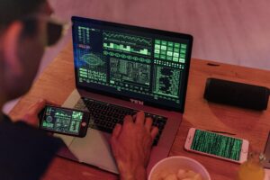 An image of a hacker's workspace, featuring a laptop with a complex, futuristic user interface, a smartphone displaying encrypted data, and various technological tools and devices.