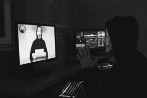 Shadowy figure in a concealing hoodie, focusing on a computer monitor showing a recognizable symbol of online anonymity.