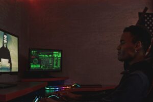 The profile of a person sitting at a desk, their face obscured as they interact with a computer displaying intricate digital information.