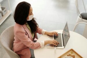 A professional-looking person wearing a mask and typing on a laptop in an office setting.