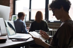 Coworkers collaborating and using technology in a shared workspace with large windows providing natural light.