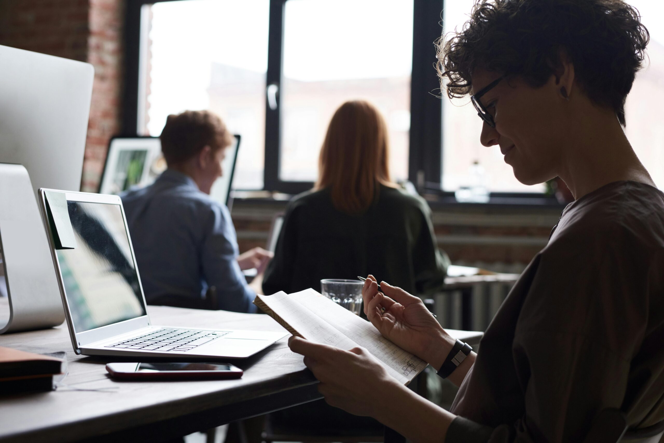 Coworkers collaborating and using technology in a shared workspace with large windows providing natural light.