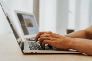 Hands typing on a laptop keyboard, with a framed photo in the background, suggesting a work or creative environment.