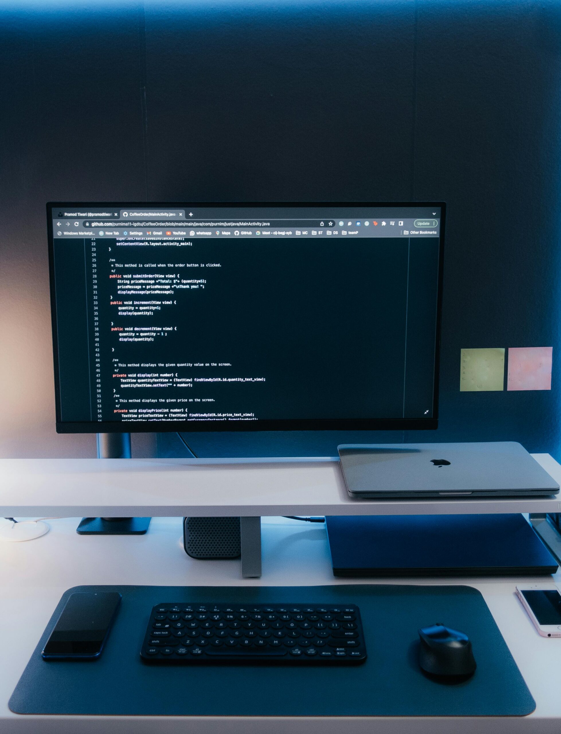 Workspace setup featuring a desktop computer monitor displaying coding, a MacBook, a wireless keyboard and mouse, and other digital accessories on a blue desk surface.