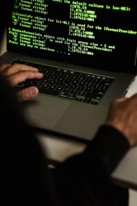 A close-up view of a person's hands typing on a laptop keyboard, with a computer screen displaying technical programming code in the background.