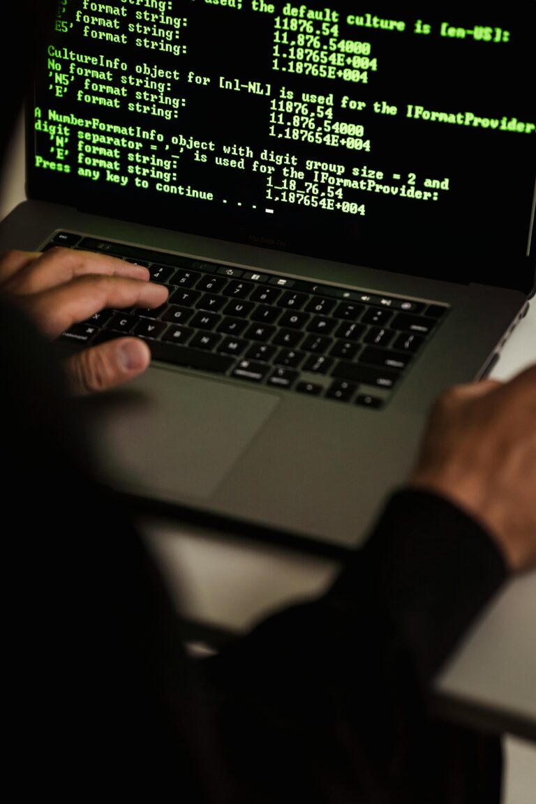 A close-up view of a person's hands typing on a laptop keyboard, with a computer screen displaying technical programming code in the background.