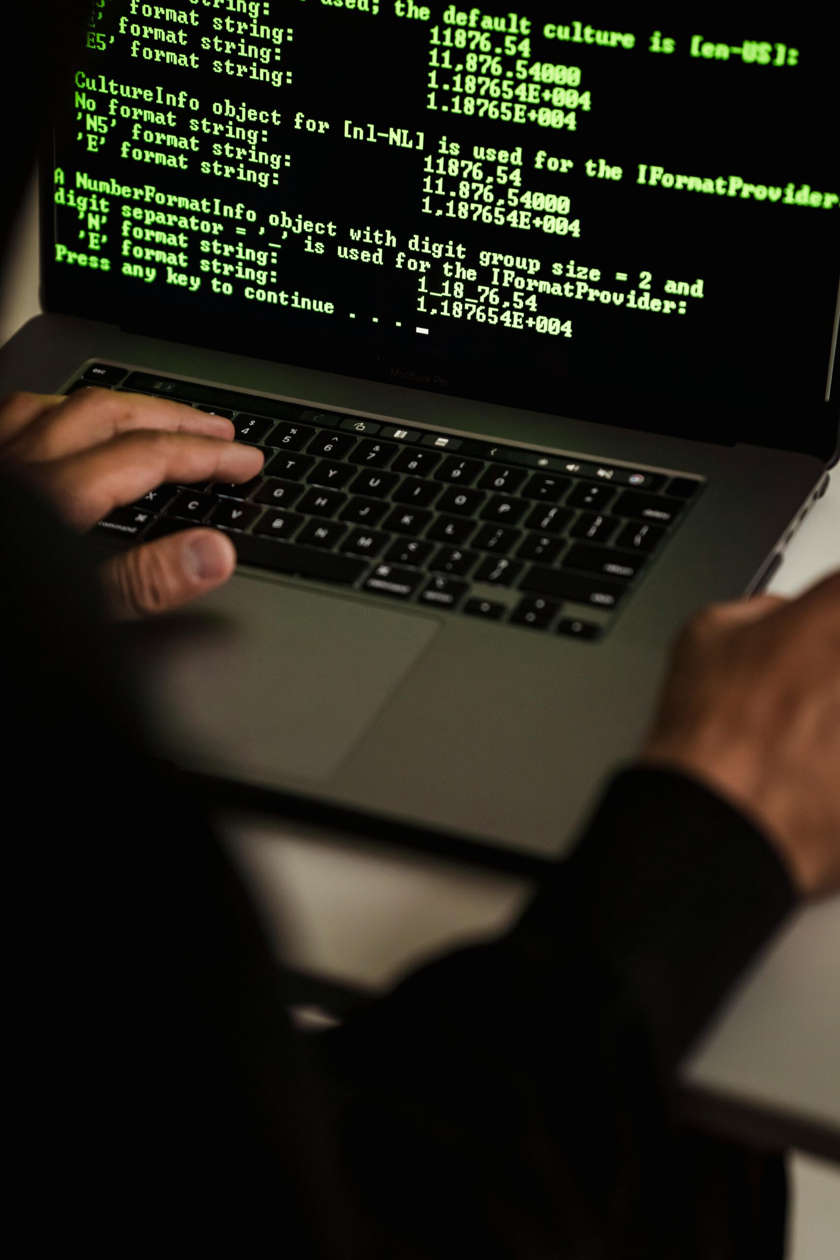 A close-up view of a person's hands typing on a laptop keyboard, with a computer screen displaying technical programming code in the background.