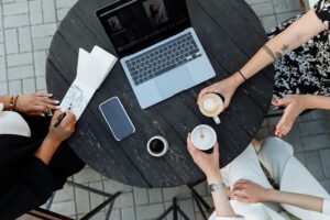 Creative workspace: A laptop, smartphone, coffee cups, and a sketchbook on a rustic wooden table, suggesting a collaborative working environment.